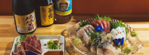 Assorted sashimi on a glass platter with wasabi, greens, and decorative flowers; sake bottles in the background.