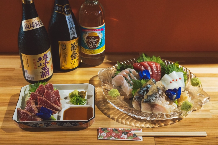 Assorted sashimi on a glass platter with wasabi, greens, and decorative flowers; sake bottles in the background.