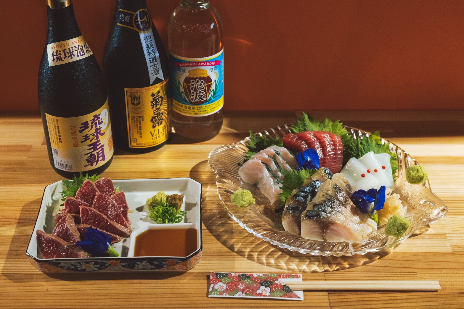 Assorted sashimi on a glass platter with wasabi, greens, and decorative flowers; sake bottles in the background.