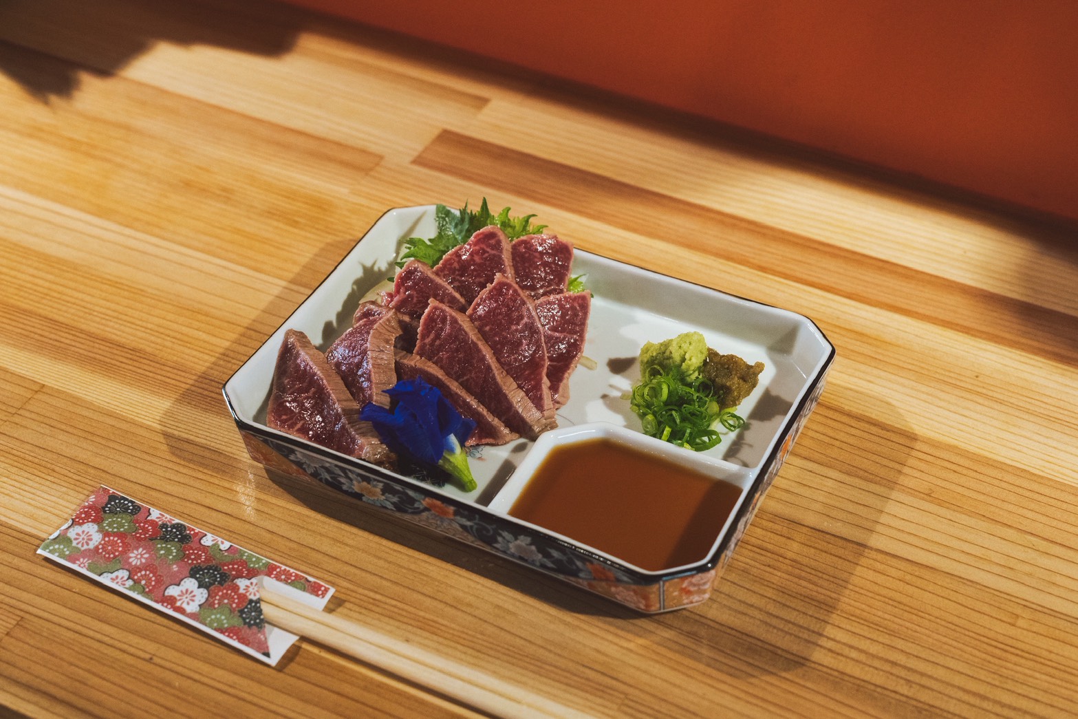 Plate of sliced tuna sashimi with soy sauce, wasabi, greens, and chives on a decorative ceramic tray