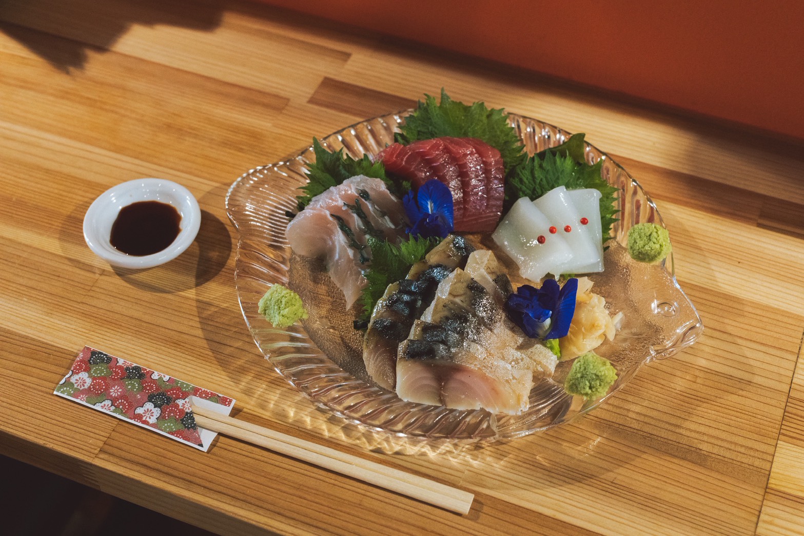 Assorted sashimi on a glass plate with greens, wasabi, and pickled ginger; soy sauce in a small dish nearby.