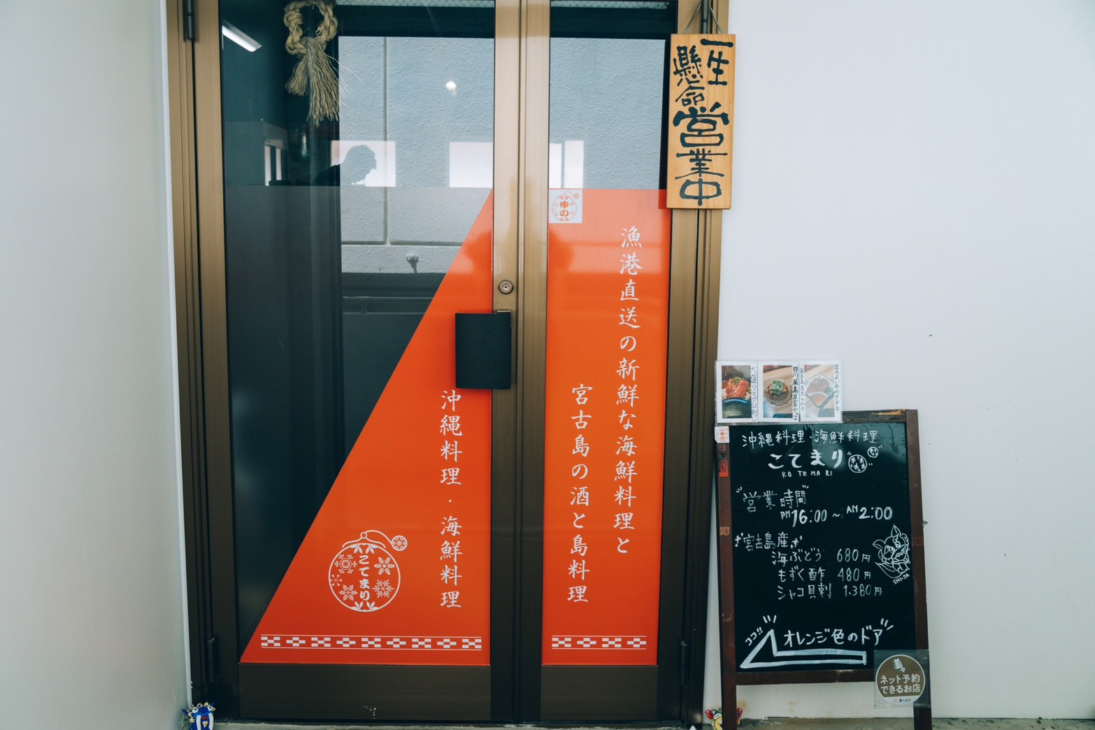 Entrance to a Japanese restaurant with tall orange vertical banners displaying white Japanese text beside glass doors and a chalkboard menu outside.
