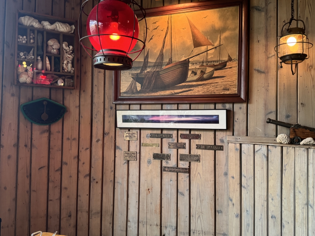 Wood-paneled wall with a framed nautical painting of sailboats, a glowing red lantern, and a shell-filled display cabinet to the left.