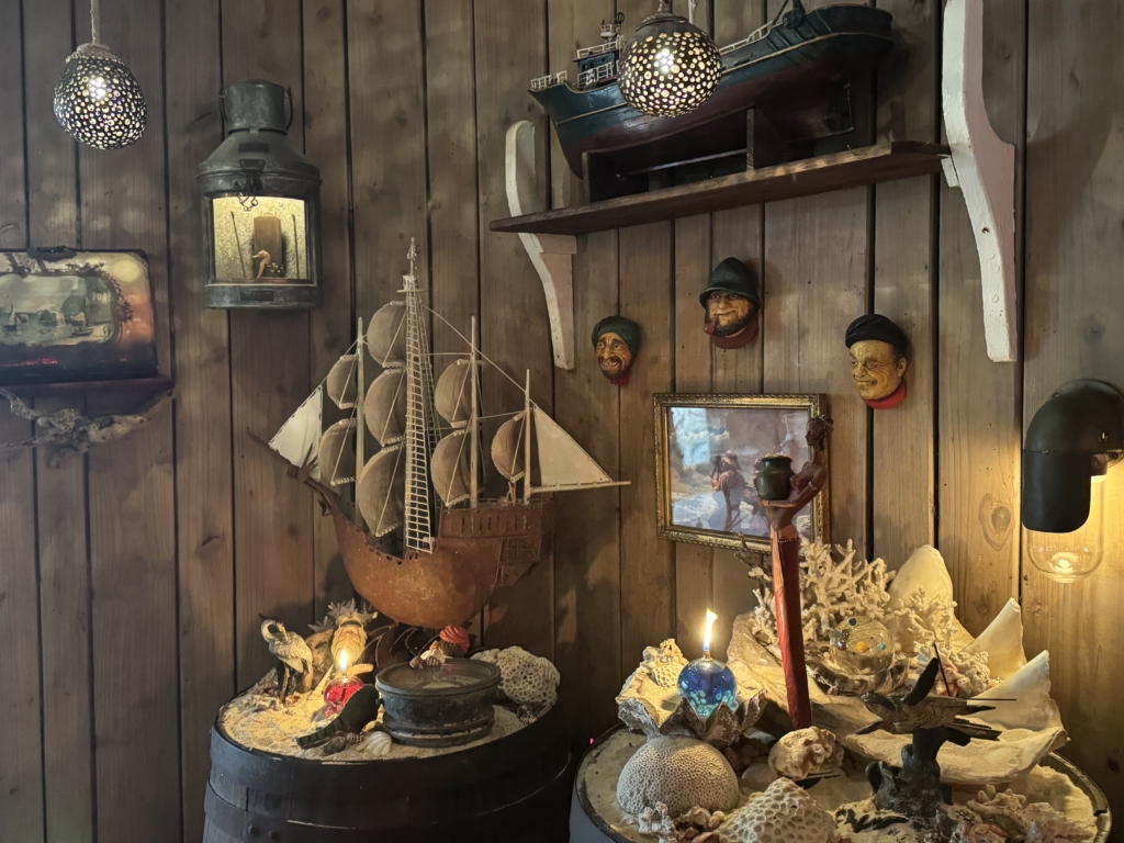 Nautical-themed display on a wooden wall: model sailboat, old lantern, carved masks, framed photo, shells, and coral accents with candles.