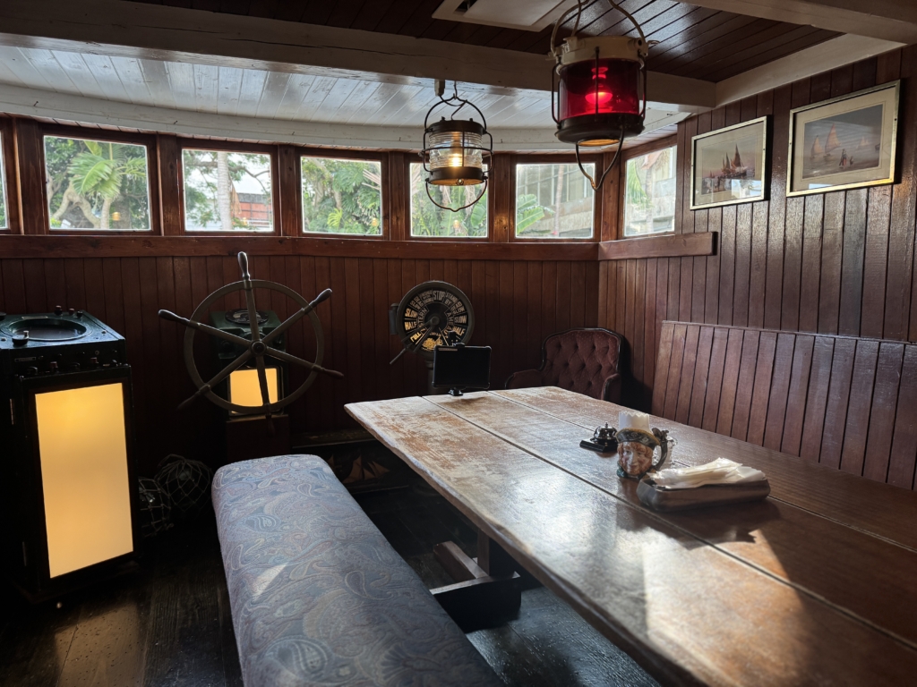 Wood-paneled nautical dining nook with a long weathered table, blue upholstered bench, ship wheel, and red hanging lanterns near windows.