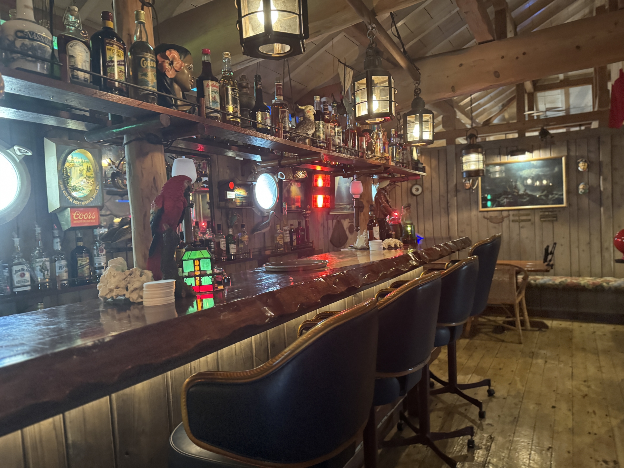 Interior of a rustic bar with a long wooden counter and shelves full of liquor bottles under hanging lanterns.