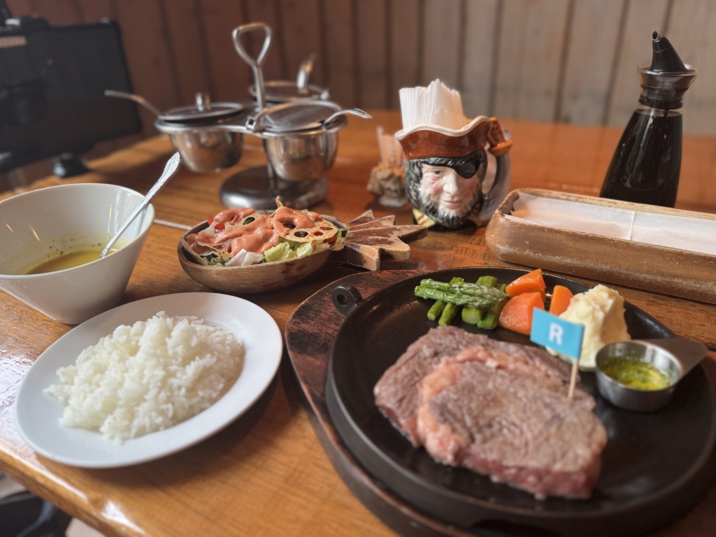 Table setting with a skillet of sliced steak and mixed vegetables, bowls of white rice, and condiments on a wooden table; a pirate-head mug in the background.