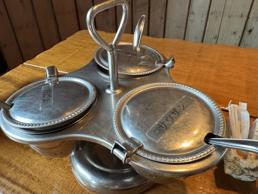 Silver three-dish serving set with beaded rims on a wooden table, central handle and lids suggesting a condiment/server tray