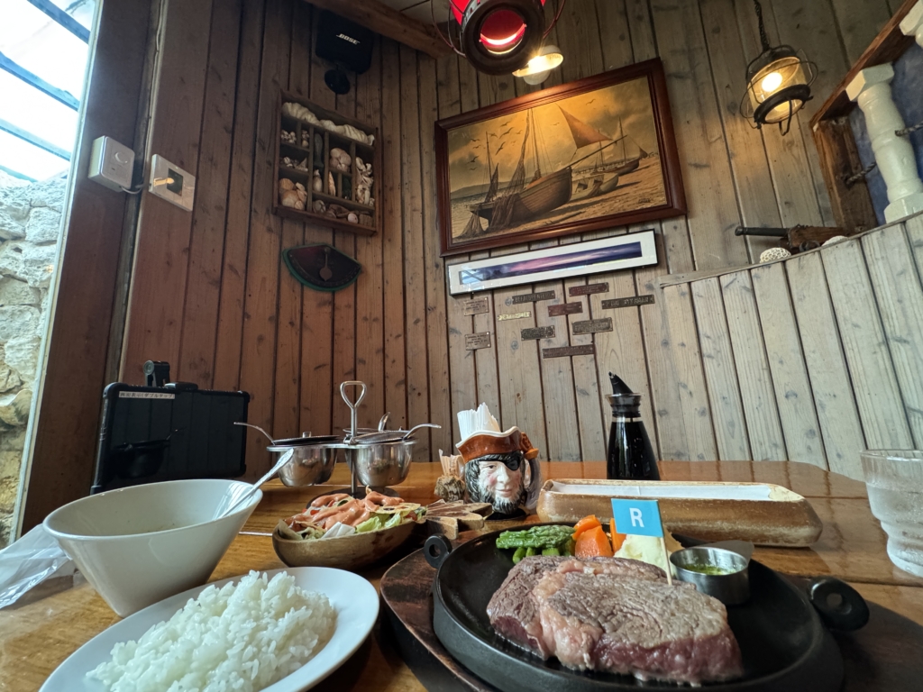 Table setup with sizzling steak on a cast-iron skillet, bowls of white rice, vegetables, and bread in a rustic wooden dining room with nautical decor and artwork in the background