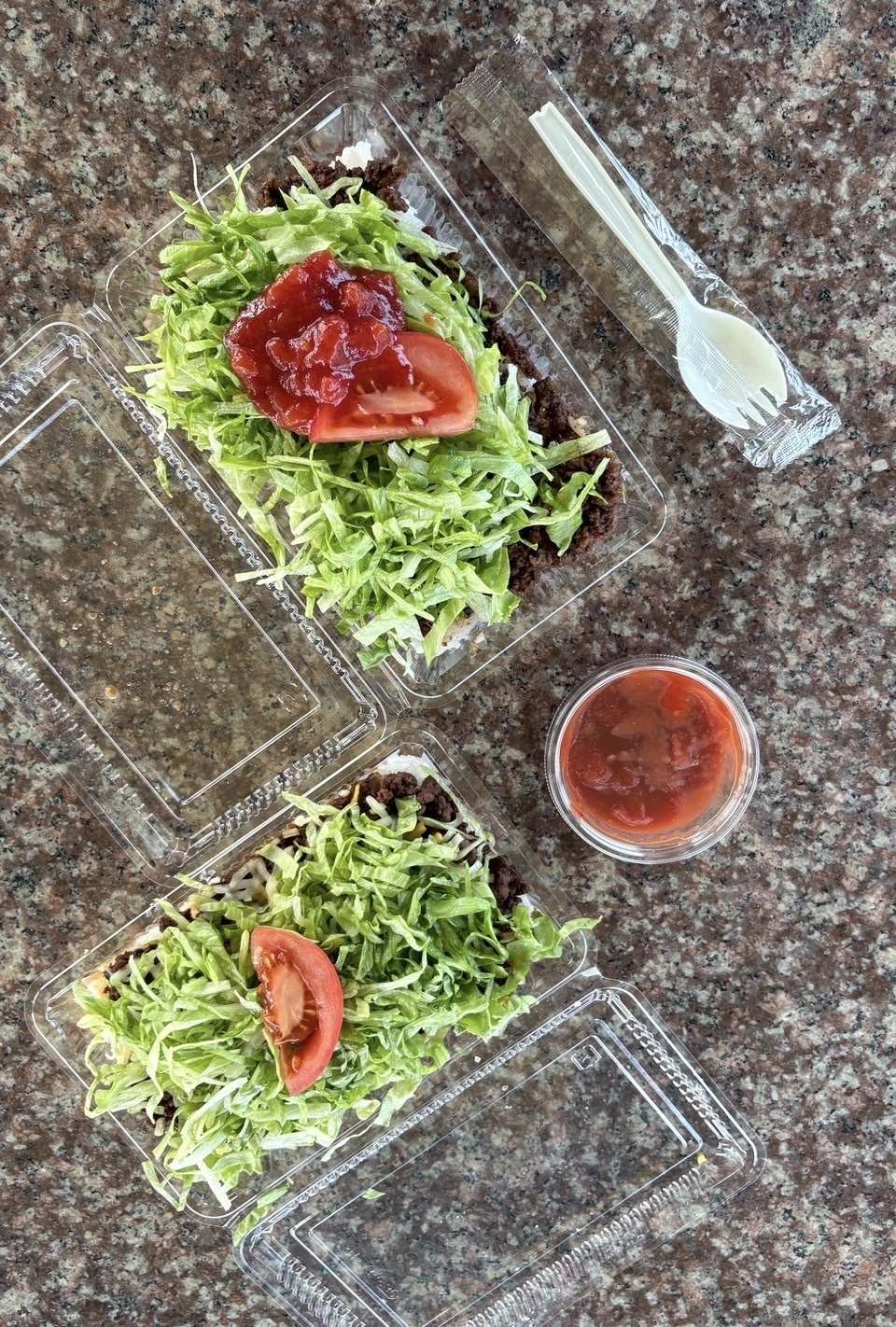 Two clear takeout salad containers filled with shredded lettuce, tomato slices, and ground beef, with a cup of red sauce on a granite counter and a plastic fork nearby.