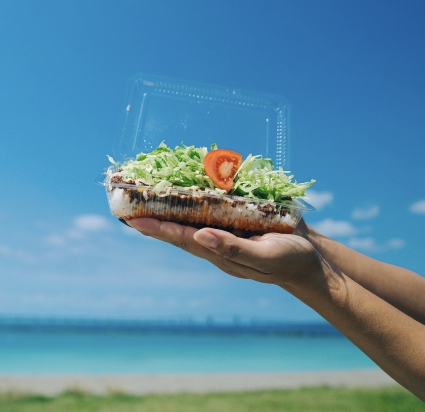 A person holds a clear plastic takeout container with a layered salad (lettuce and tomato) against a blue seaside sky.